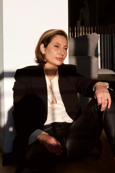Furniture designer Christina Z Antonio wearing a black suit and slightly open white button-down while sitting in a chair.