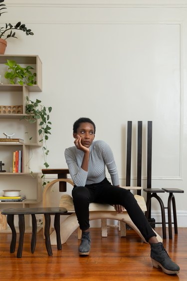 Furniture designer Nifemi Ogunro sitting on a piece of wood furniture, wearing black pants, black suede cloglike boots, and a long light gray T-shirt.