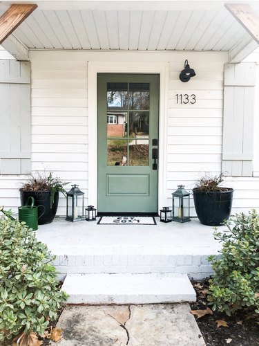 A green door on a house with white siding, black modern house numbers, and a black porch light