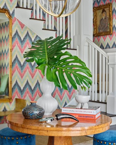 Tablescape with monstera fronds in white vase, stacked books, a horned magnifying glass, and other decorative objects.
