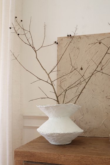 DIY paper mache vase with dried branches on cabinet in front of textured canvas painting