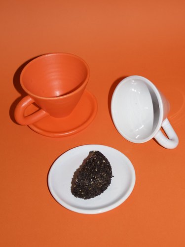 An orange mug with a saucer next to a fallen white mug topped with coffee grounds on an orange background.