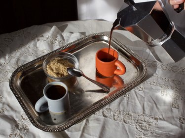 An orange mug, a white mug, and a bowl with a spoon on a metal tray. The tray is resting on a white surface, and there is a hand pouring coffee into the orange mug.