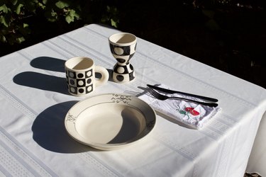 A bowl, two cream-colored cups, and silverware on a table with a white tablecloth.
