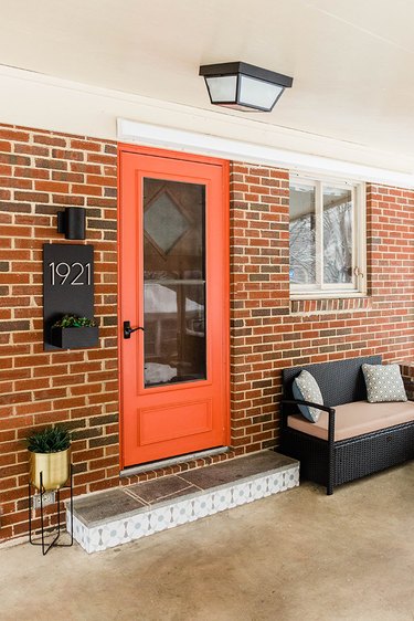Brick house with an orange storm door, gold plant stand, black sconce light, and black bench