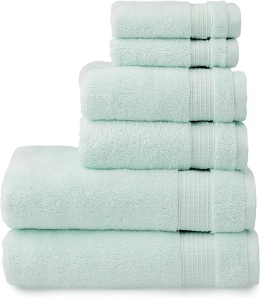 A stack of two turquoise bath towels, two turquoise hand towels, and two turquoise washcloths rest on top of one another in front of a white background.