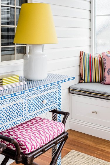 Room detail with blue and white bone inlay table, white lamp with bright yellow lampshade, dark-wood stool with hot pink and white print cushion, and built-in bench.