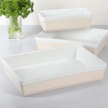 Three light pink and white stoneware baking dishes (9-inch, 11-inch and 13-inch) rest on a white kitchen counter, with a glass of herbs nearby.
