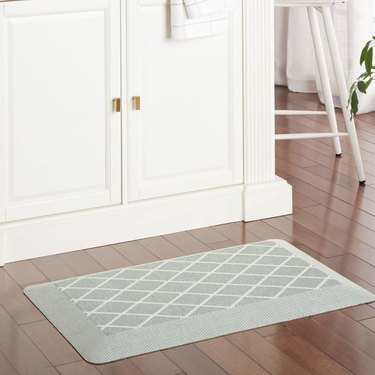 A mint green kitchen mat with a white diamond design rests on a dark wood kitchen floor in front of two white cabinet doors with gold handles.