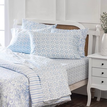 A white bed with light blue medallion pillows, sheets and comforter. A white bedside table is to the side, with a white vase and greenery.