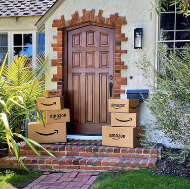 Seven Amazon boxes stacked on top of one another rest outside a home with a dark wood door with a black handle. The home is surrounded by greenery, and a brick walkway, with two brick steps lead to the front door. Four Amazon boxes are stacked to the left and three are stacked on the right.