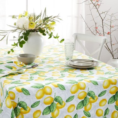 A dining table with a tablecloth featuring bunches of lemons. A white of vase of yellow flowers and greenery rests on the table, along with a neutral colored place setting, with a glass cup. A bowl also rests on the table, with a lemon napkin and spoon.