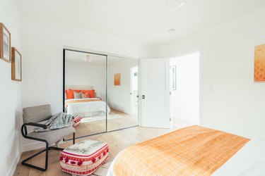 Bedroom with white bedding with orange pillows, light wood floor, mirrored closet, and gray armchair