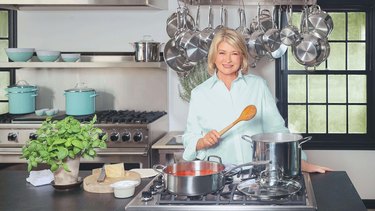Martha Stewart stands in front of a stove in a large kitchen, holding a wooden spoon. She is wearing a light green button up shirt, and two large pots rest on the stove in front of her. There is a block of cheese and a basil plant to her side, and behind her is a window, rows of hanging pots and pans, and two turquoise pots resting on another stove. There is a shelf above the stove with three turquoise and white bowls, along with another silver pot.