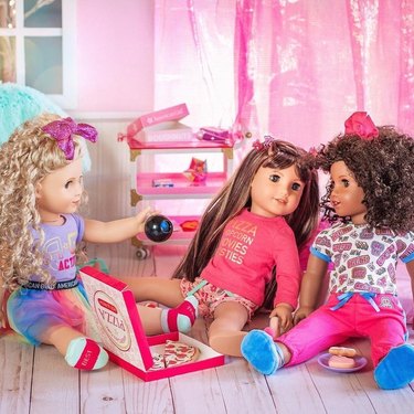 Three American Girl dolls sitting on the floor of a pink bedroom next to a box of pizza