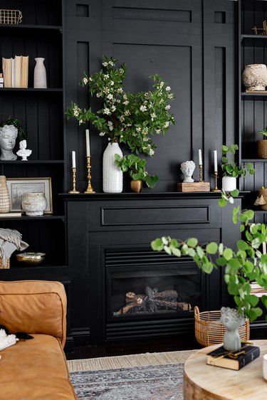 Black fireplace mantel and shelves styled with white busts, brass candlesticks, wood elements, and greenery.