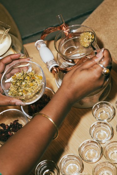 A hand scooping herbs into a clear glass canister.