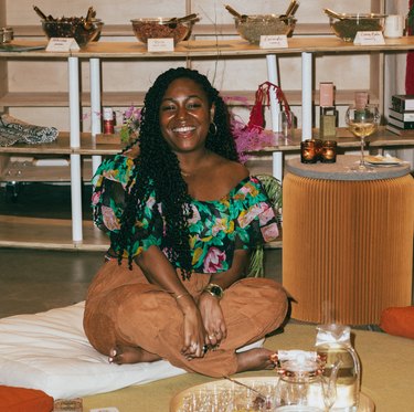 Branché Foston wearing a floral green top and brown pants sitting cross-legged on the floor next to a tray of herbs and tea.