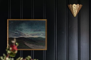 Black paneled wall with a gold scalloped Art Deco sconce and a landscape painting.