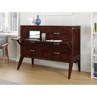 A dark wood cabinet bed that is shown in the form of a desk with six drawers and a pulled-down work surface with a laptop and papers on it.