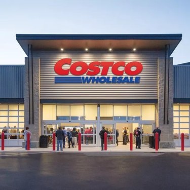A Costco warehouse at night. The red and blue logo is on the front of the building, with four lights above it. There is a line of people outside the doors, spilling onto the sidewalk.