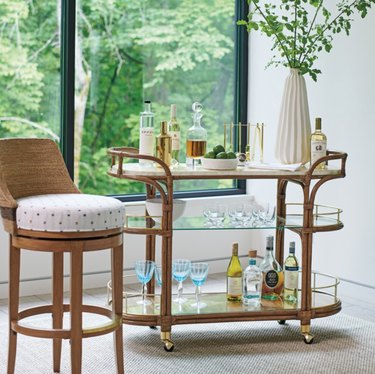 Bamboo and glass bar cart with vase, glassware, bottles, barstool.