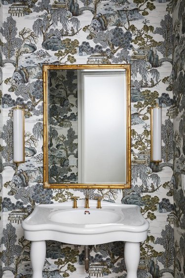Powder bathroom with gray and gold chinoiserie wallpaper, white sink, gold mirror, and light bar sconces