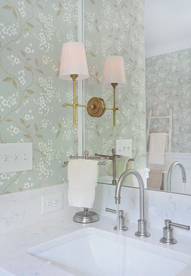 Bathroom vanity with pale blue-green floral wallpaper and mirror sconce in brass with white lampshade