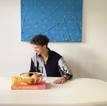 Artist Werner Bronkhorst sits at a white kitchen table and is looking to the side and smiling. He is a white man with curly short brown hair and is wearing a white and gray long-sleeve button-up top. A bright pink book with "Palm Springs" in bright yellow on the spine rests on the table with a bowl of oranges and a yellow glass on top. A large royal blue canvas with various miniature surfers painted throughout is behind him, titled "Board Meeting"