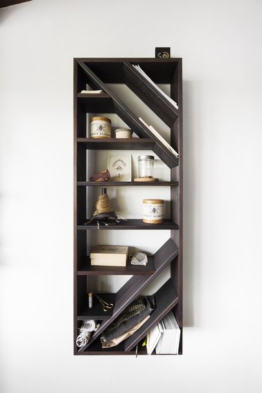 A dark wood vertical bookshelf on a white wall with off-kilter shelving holding small objects and papers.