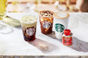 Three Starbucks drinks next to a small canister of Partanna olive oil on a white surface.