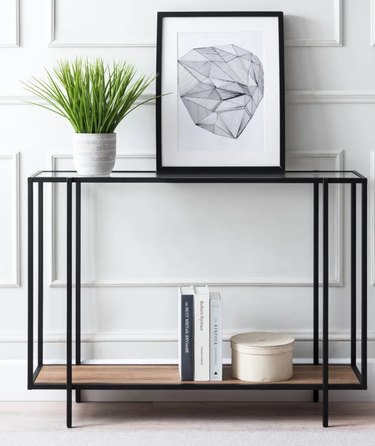 Black metal console table with glass and wood shelves, plant, books, art.
