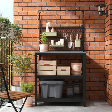 A black stainless steel outdoor kitchen island against a red brick wall on a balcony, featuring kitchen cooking tools and a potted tomato plant.