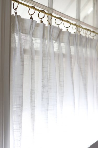 No-sew white pinch pleat cafe curtain hanging in window with brass curtain rod