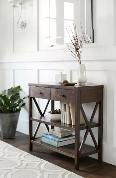 Dark brown wood console table with drawers and shelf, vase, books, mirror, rug.