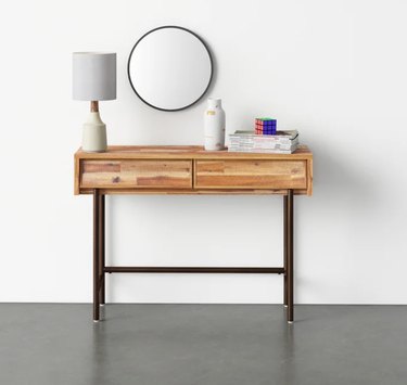 Light wood and metal console table with drawers, lamp, mirror, books, vase.