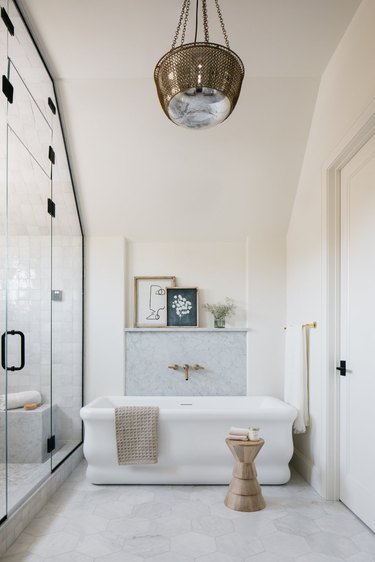 White and gray bathroom with large white bathtub with picture ledge behind it and brass antique style chandelier