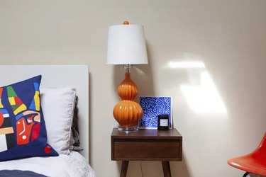 Wood modernist style night table, with an orange table lamp, and a blue-white print. A bed with a colorful, abstract pillow.