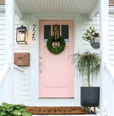 A pink front door on a white house; a black lantern and black modern house numbers are on the left side of the door