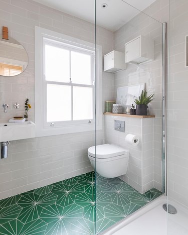 Small bathroom with light gray tiled walls and sunburst green tile and ledge behind the toilet with plants.