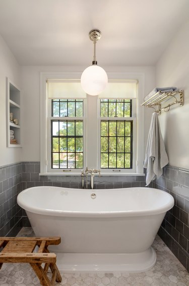 Modern traditional bath nook with large white bathtub, pendant light overhead, black framed windows behind, and different kinds of shelving on either side