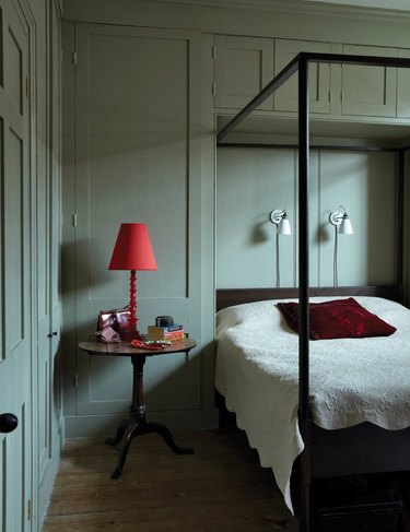 Light green-grayish painted bedroom with black canopy bed and wood side table with red lamp