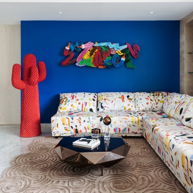 Red cactus coat stand against blue accent wall in living room with colorful confetti-patterned white sectional on tan carpet