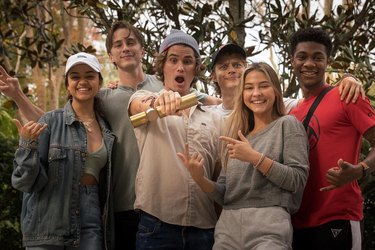 The cast of 'Outer Banks' on Netflix laughing and smiling in front of trees.