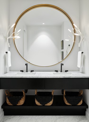 Black, white, and brass bathroom with statement sconces, brass mirror, and black and wood-toned baskets