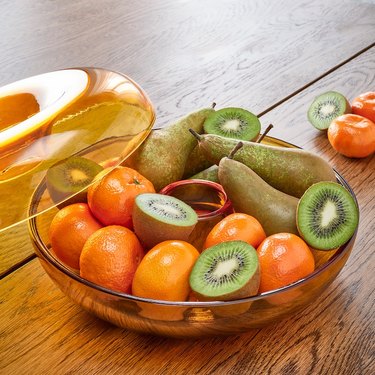 IKEA's VARMBLIXT serving bowl. The bowl rests on a wooden surface and is filled with clementines, halved kiwis and pears. The lid is resting to the side of the bowl..