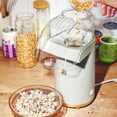 A white popcorn maker next to a bowl of popcorn on a wooden countertop.
