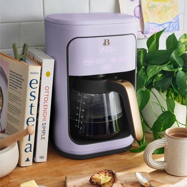 A lavender coffee maker on a wooden counter next to books and plants.