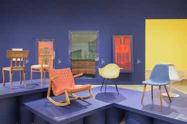 photo showing a museum installation with blue walls, chairs on pedestals, and weavings on the wall