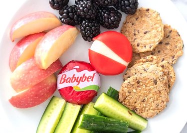 Two Babybel cheese wheels on a white plate next to slices of red apple, blackberries, cucumber, and crackers.